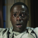 Close-up of a distressed Black man with wide eyes and a tear streaming down his cheek, sitting in a dimly lit room, appearing hypnotized and frozen in fear.
